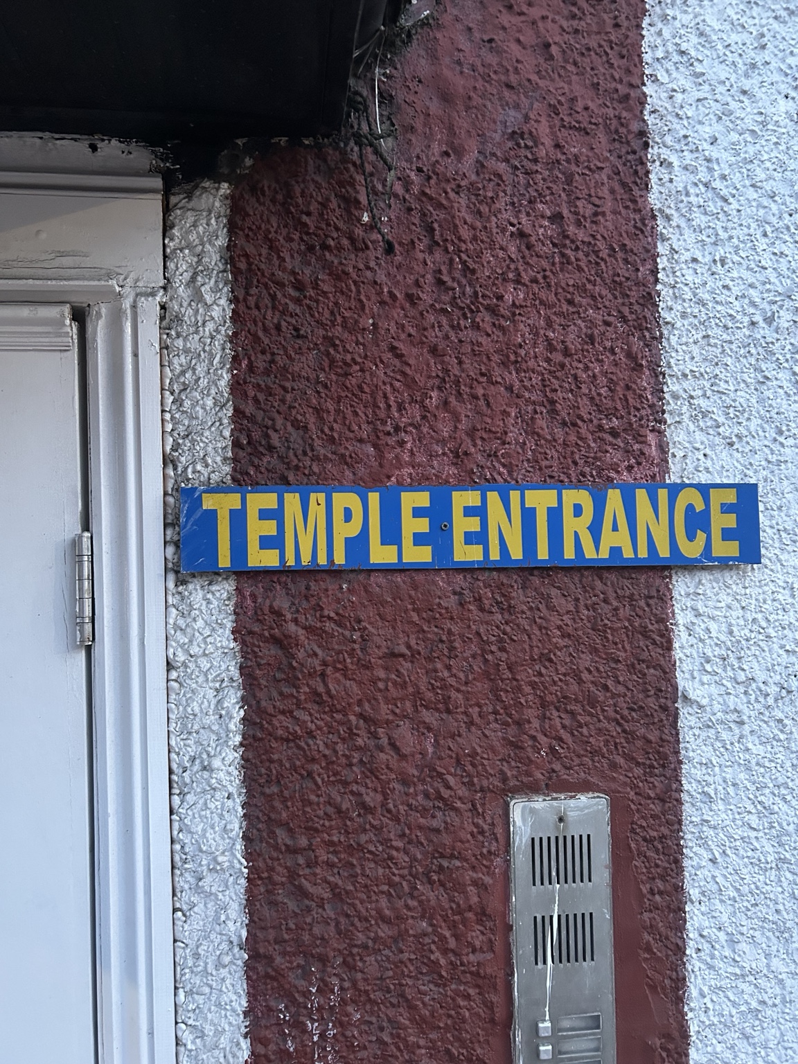A blue and yellow Temple Entrance sign beside a doorway at London Sree Ayyappan Temple.